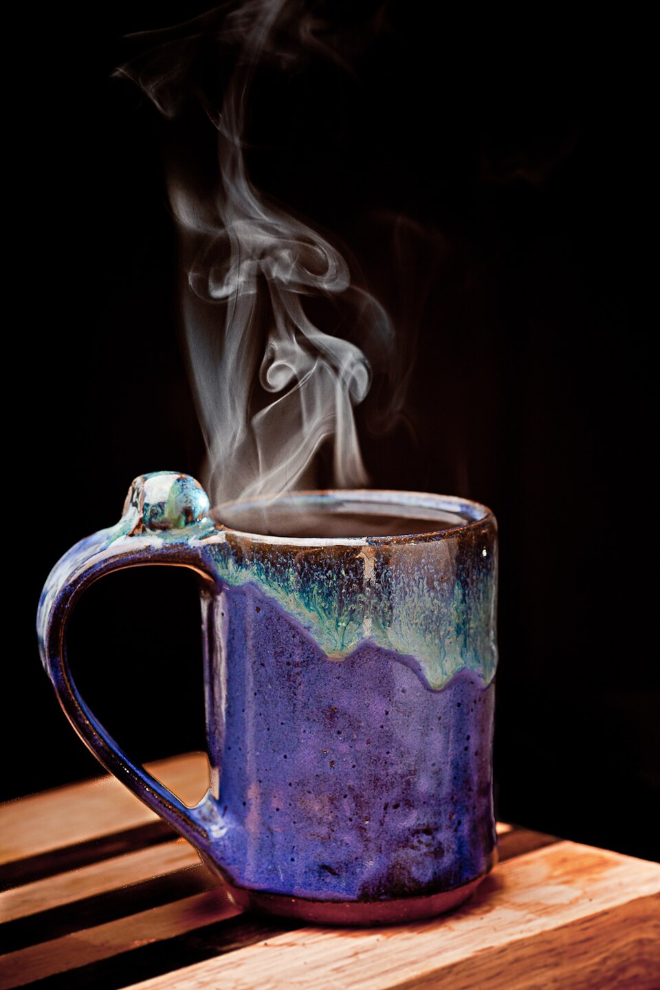 Steaming handmade stoneware mug with a blue and green drip glaze on a wooden board against a black background