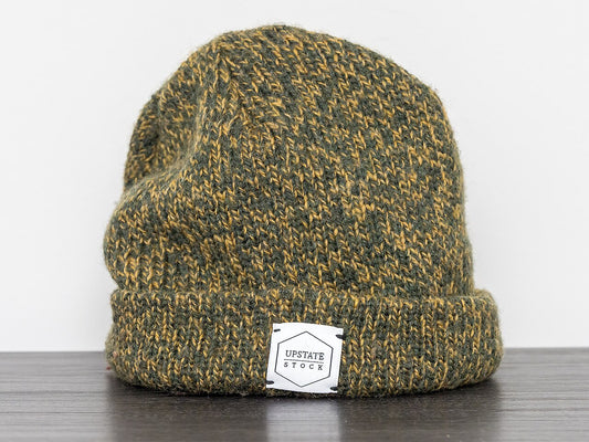 Green and gold marled ribbed knit beanie with a folded cuff, standing on a dark wood surface