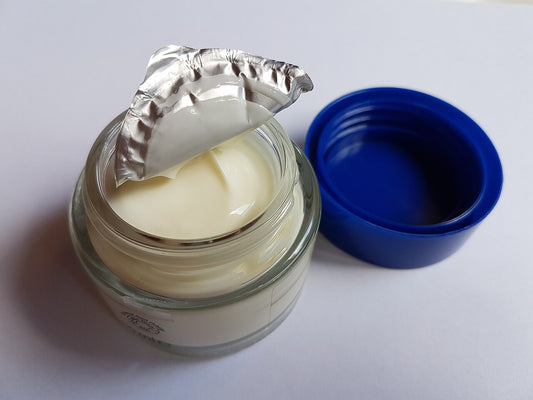 Open glass jar of thick white cream with the foil seal peeled back and a blue screw lid resting beside it