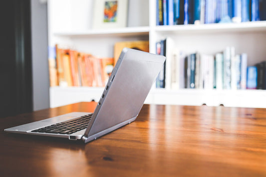 An open silver laptop on a wooden table with a blurred white bookshelf behind it