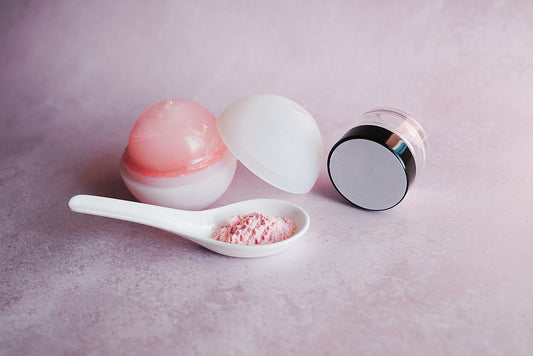 Open round cosmetic jar with its lid off beside a small ceramic spoon of pink powder on a pink surface