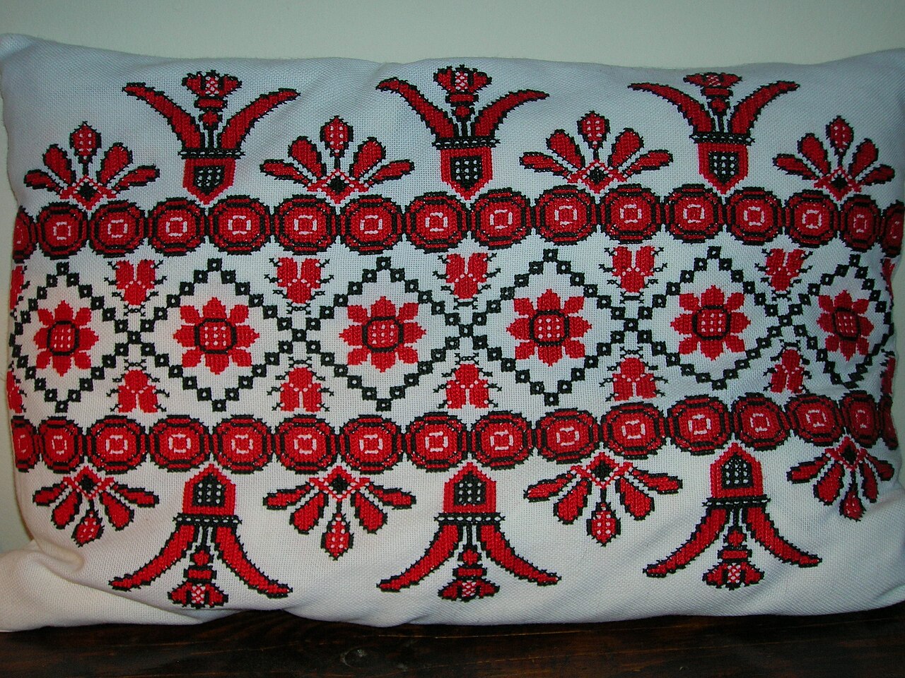 Rectangular white fabric cushion covered in red and black cross-stitch floral embroidery, resting on a dark wood surface