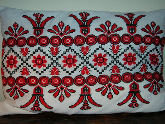 Rectangular white fabric cushion covered in red and black cross-stitch floral embroidery, resting on a dark wood surface