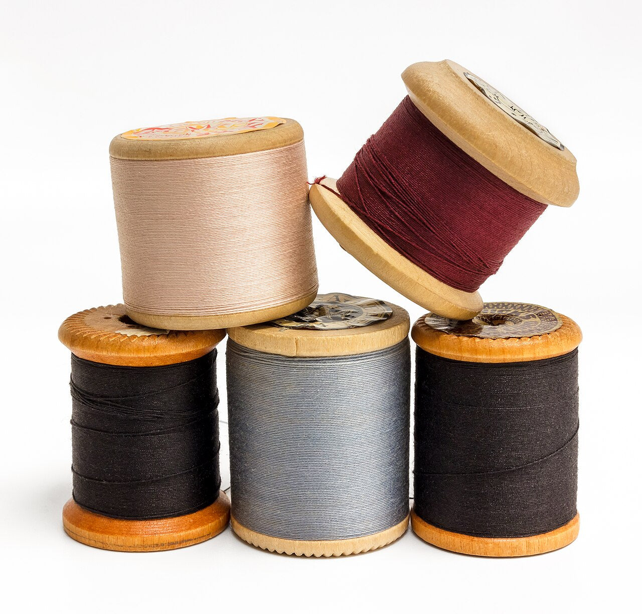 Five wooden spools of sewing thread in cream, dark red, grey and black stacked on white
