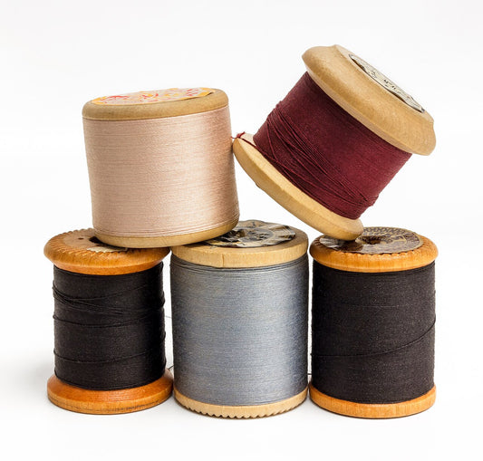 Five wooden spools of sewing thread in cream, dark red, grey and black stacked on white